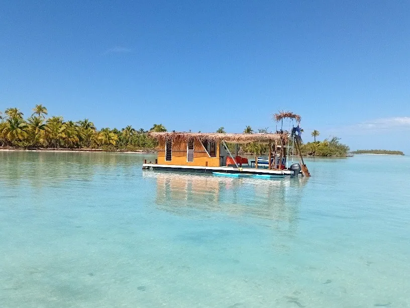 A small open-sided floating lounge on clear Aitutaki Lagoon water, with a palm-lined shore behind it.